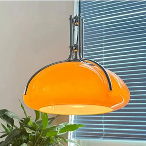 The orange glass shade lit in front of a window blind beside a green plant