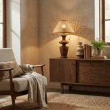 Rattan table lamp lit on a wooden sideboard beside books and a plant