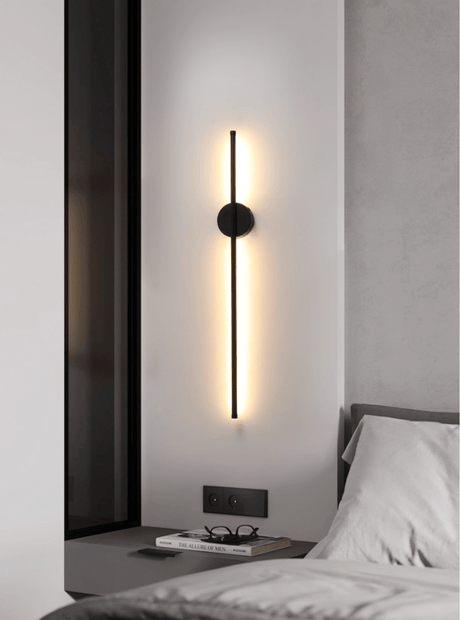Mounted vertically beside a bed, the bar lit against a pale bedroom wall