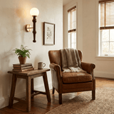 Sconce lit above a plant on a wood stool beside a wing chair and a window