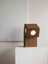 The smaller teak lamp lit on a white plinth against a pale wall
