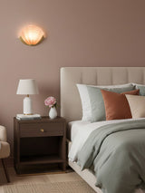 A shell sconce lit above a nightstand and a bed on a dusty pink wall