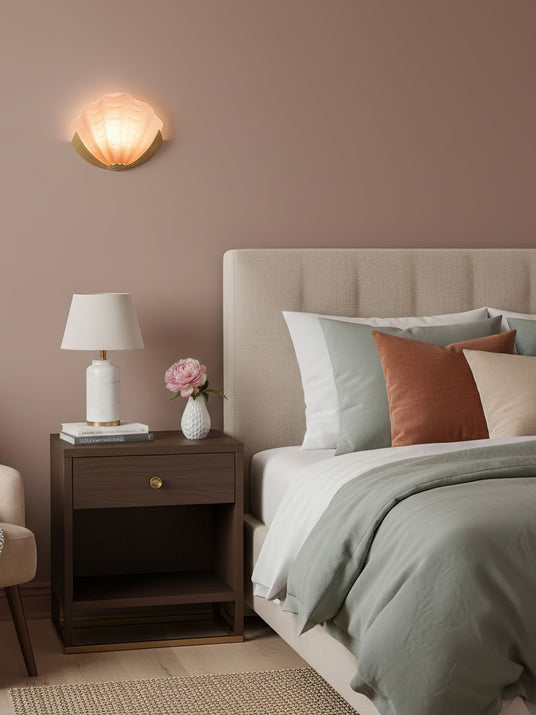 A shell sconce lit above a nightstand and a bed on a dusty pink wall