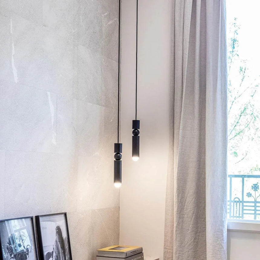 A slim gold tube pendant lit beside a long curtain and a framed picture