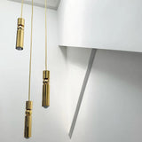 Two slim gold tube pendants lit at different heights against a pale wall