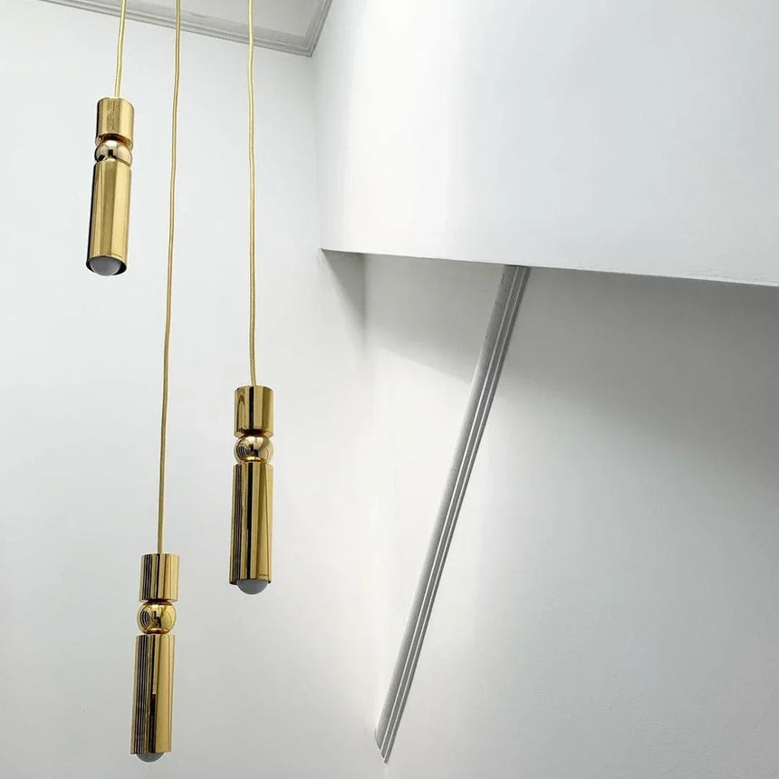 Two slim gold tube pendants lit at different heights against a pale wall