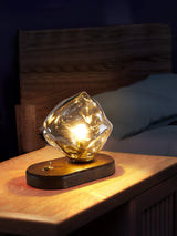 Glass ice form lamp lit on a bedside shelf against a wood wall