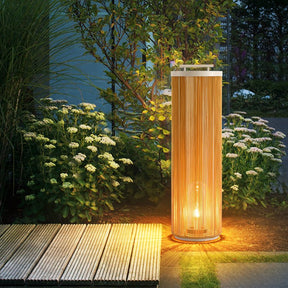 Rattan column lamp lit on decking beside white flowers in a border