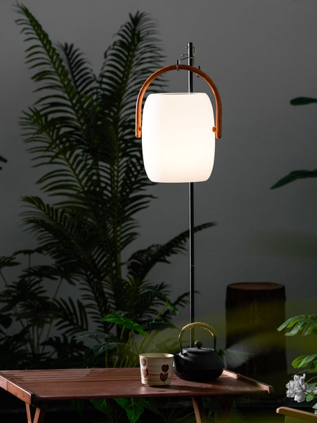 The lamp lit on a wood table with ferns and tall planting behind it