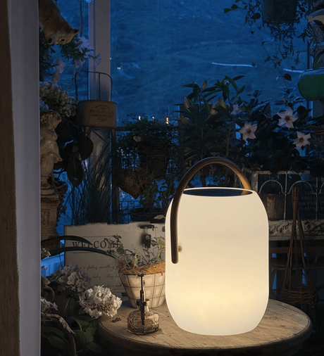 The lamp lit on a round garden table with planting and a chair behind