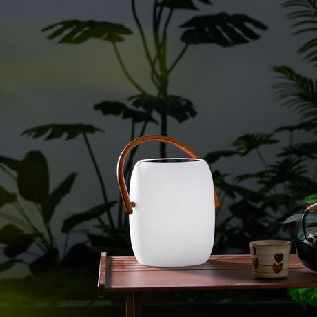 The lamp lit on a small side table with dark foliage rising behind it