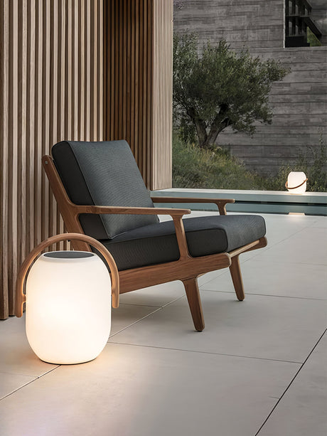 The lamp lit on paving beside a timber armchair at the edge of a pool