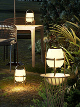 The lamp lit on a garden table with chairs and planting behind at dusk
