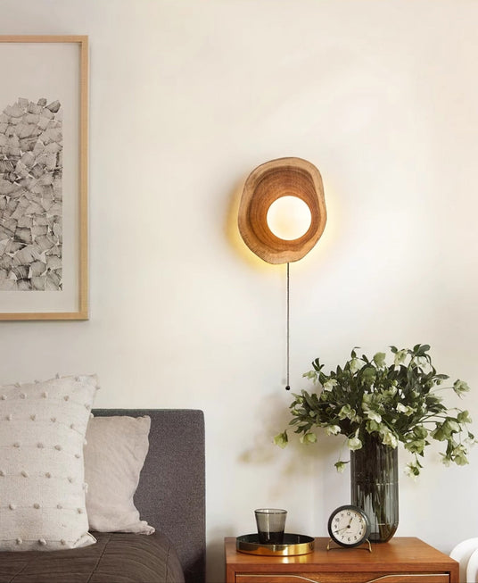Sconce lit above a nightstand with a vase of foliage and a clock by a bed