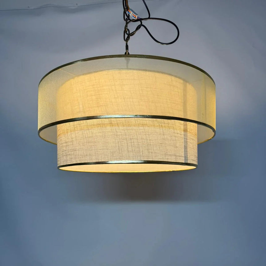 Photograph of the tiered drum pendant light lit, showing the woven beige fabric and metal trim