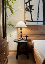 Spindle table lamp lit on a dark table beside a bed and a wood cabinet