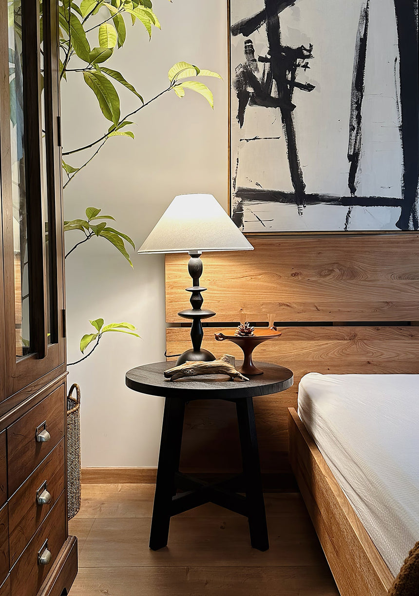 Spindle table lamp lit on a dark table beside a bed and a wood cabinet