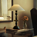 Spindle table lamp lit on a console below a framed mirror with flowers