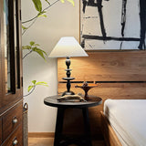 Spindle table lamp lit on a round side table beside a wood framed bed
