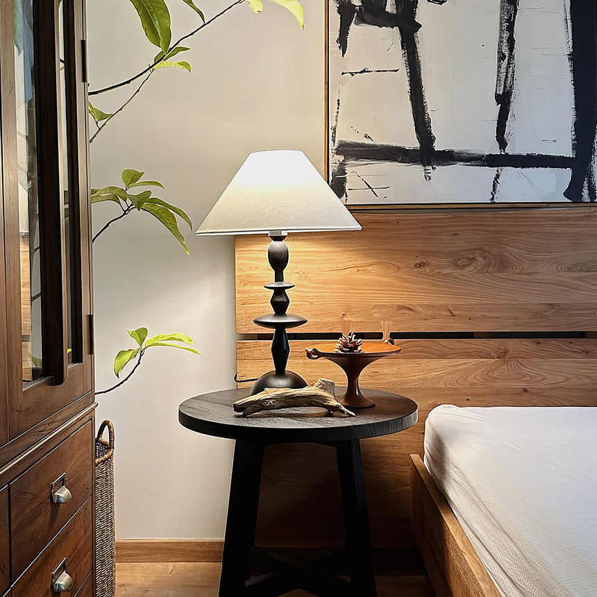 Spindle table lamp lit on a round side table beside a wood framed bed