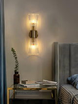 The sconce above a glass nightstand with books and a plant beside a gray headboard