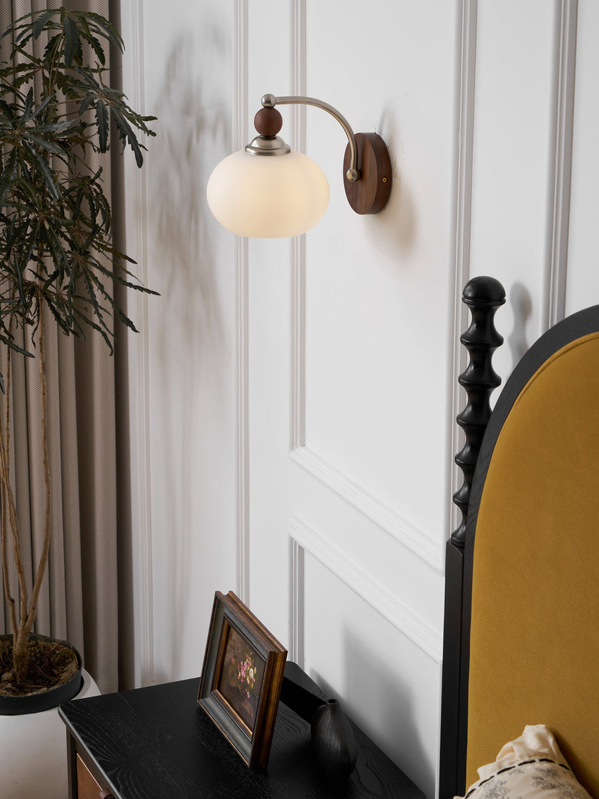 Sconce lit beside a bed with a plant leaning in over a dark nightstand