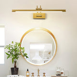 Brass bar lit above a round gold mirror on a vanity with a plant and small bottles