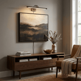 Dark bronze bar lit over a dark landscape canvas above a low walnut console and chair