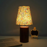Floral shade lamp on a wood cylinder base lit against a dark ground