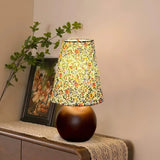 Floral shade lamp lit on a wood bench beside a framed botanical print