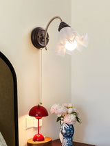 Sconce lit in a corner above a nightstand with a red lamp and a vase of flowers