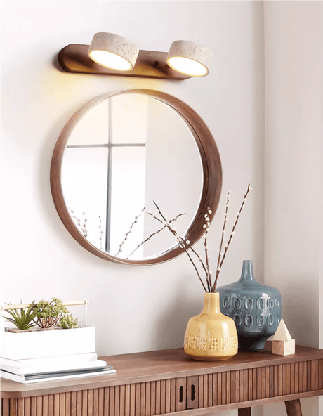 Lit above a round mirror over a sideboard holding a vase and a stack of books