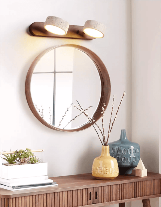 Lit above a round mirror over a sideboard holding a vase and a stack of books