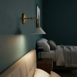 The brass sconce lit on a dark teal bedroom wall above a bed and a bench