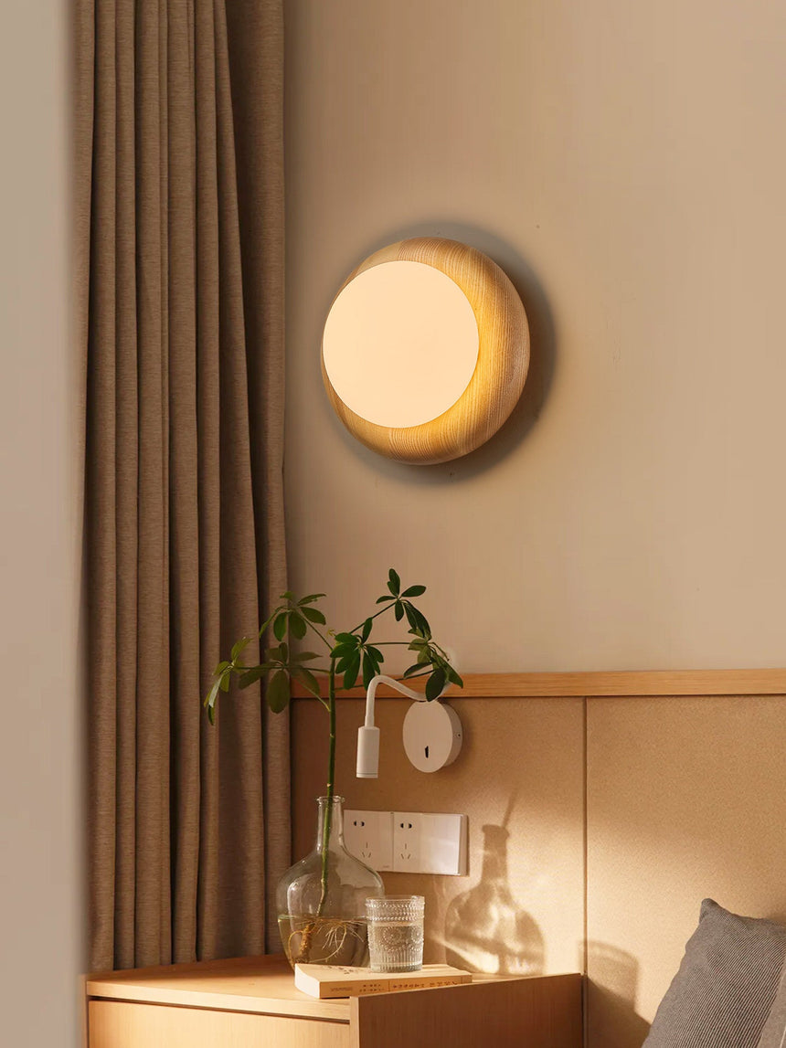 Sconce lit beside a bed above a nightstand with a glass carafe and a curtain