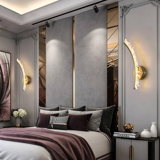 A pair of curved crystal sconces lit on each side of a bed with purple linens