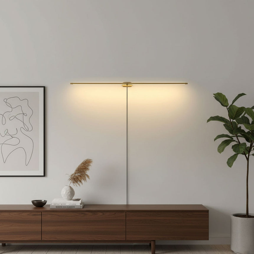 Slim gold rod lit on a pale wall between a framed print and a tall plant