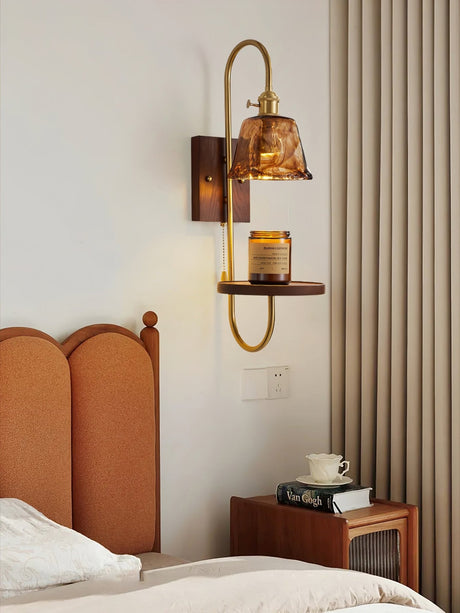 Sconce lit beside a bed, a cup and a book below the lit shade