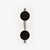 Wall light with two black discs on a polished rod, soft light rippling behind them