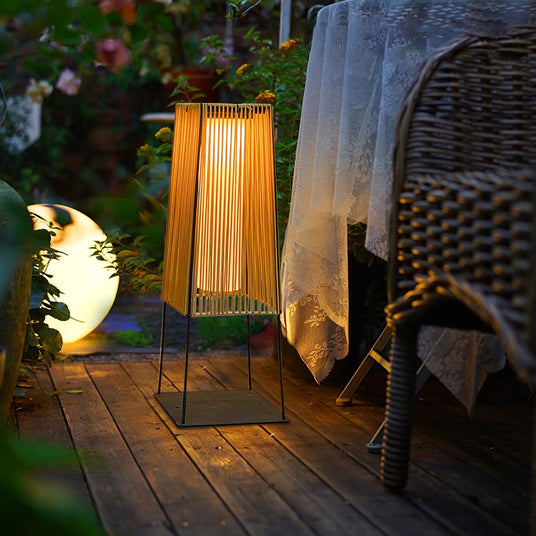 The lamp lit on a timber deck beside a woven chair and a soft throw